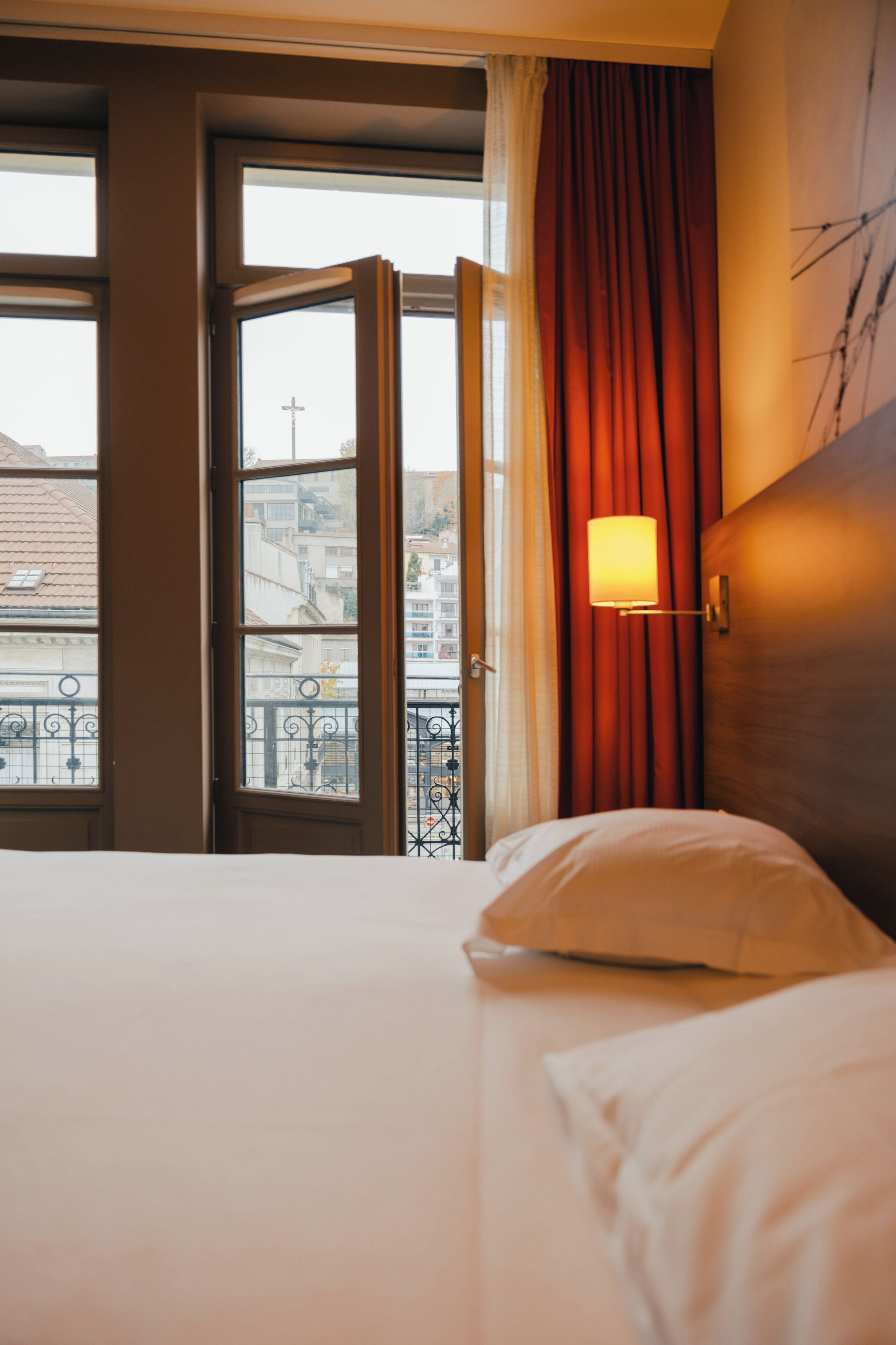 A hotel room with a bed, a window with curtains, and a view of buildings outside.