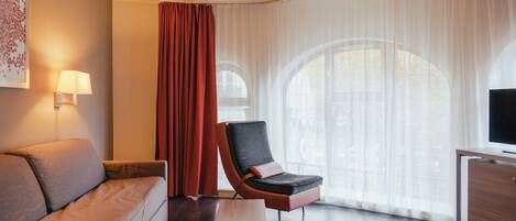 Una habitación de hotel moderna con sofá, una silla, televisor y un ventanal con cortinas.