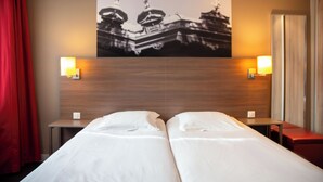 A hotel room with two beds, a wooden headboard, two bedside tables with lamps, a red curtain, and a wall-mounted picture of a building.