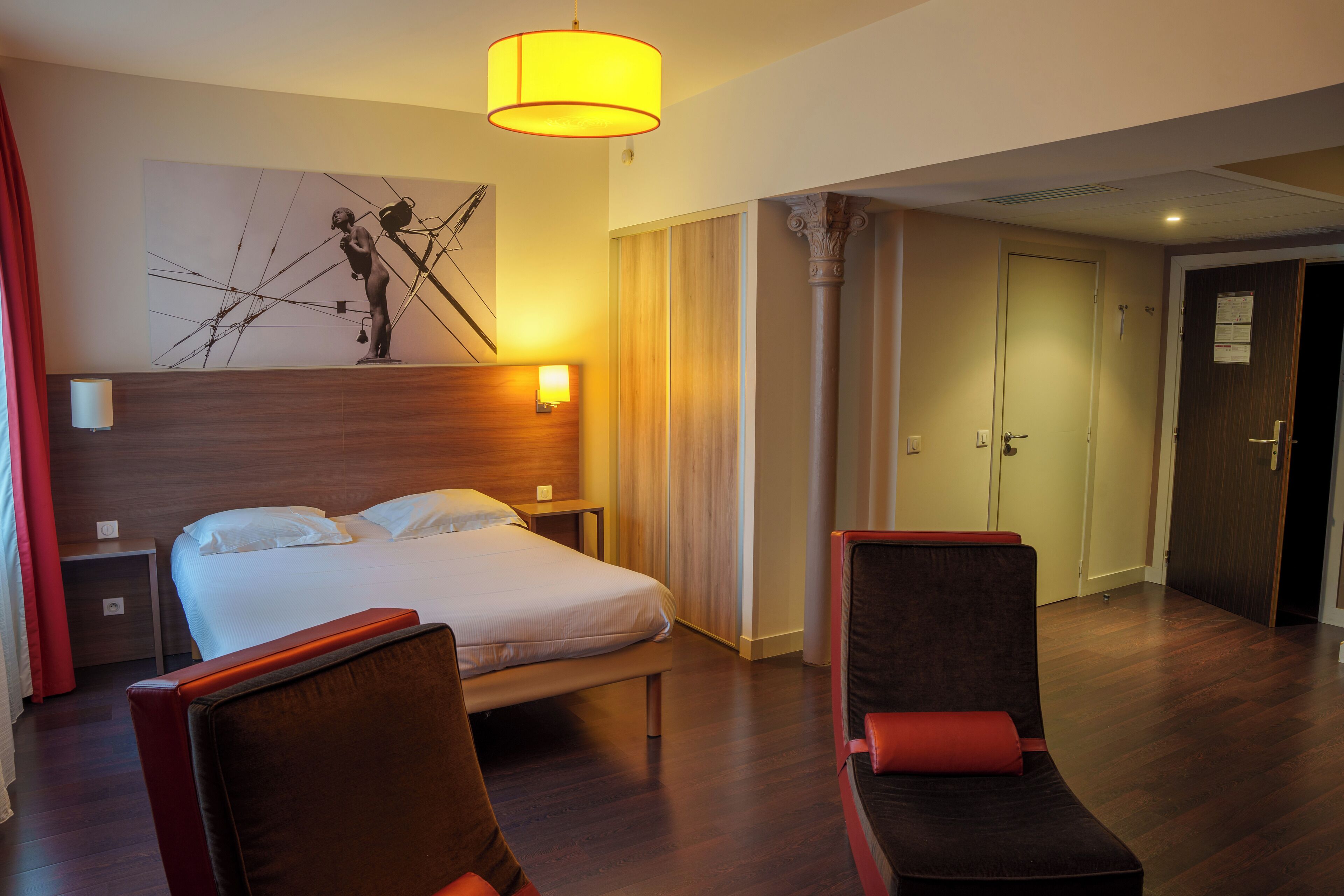 Habitación de hotel con cama, dos sillones, una mesita de noche, una lámpara de pared y un cuadro abstracto de gran tamaño.