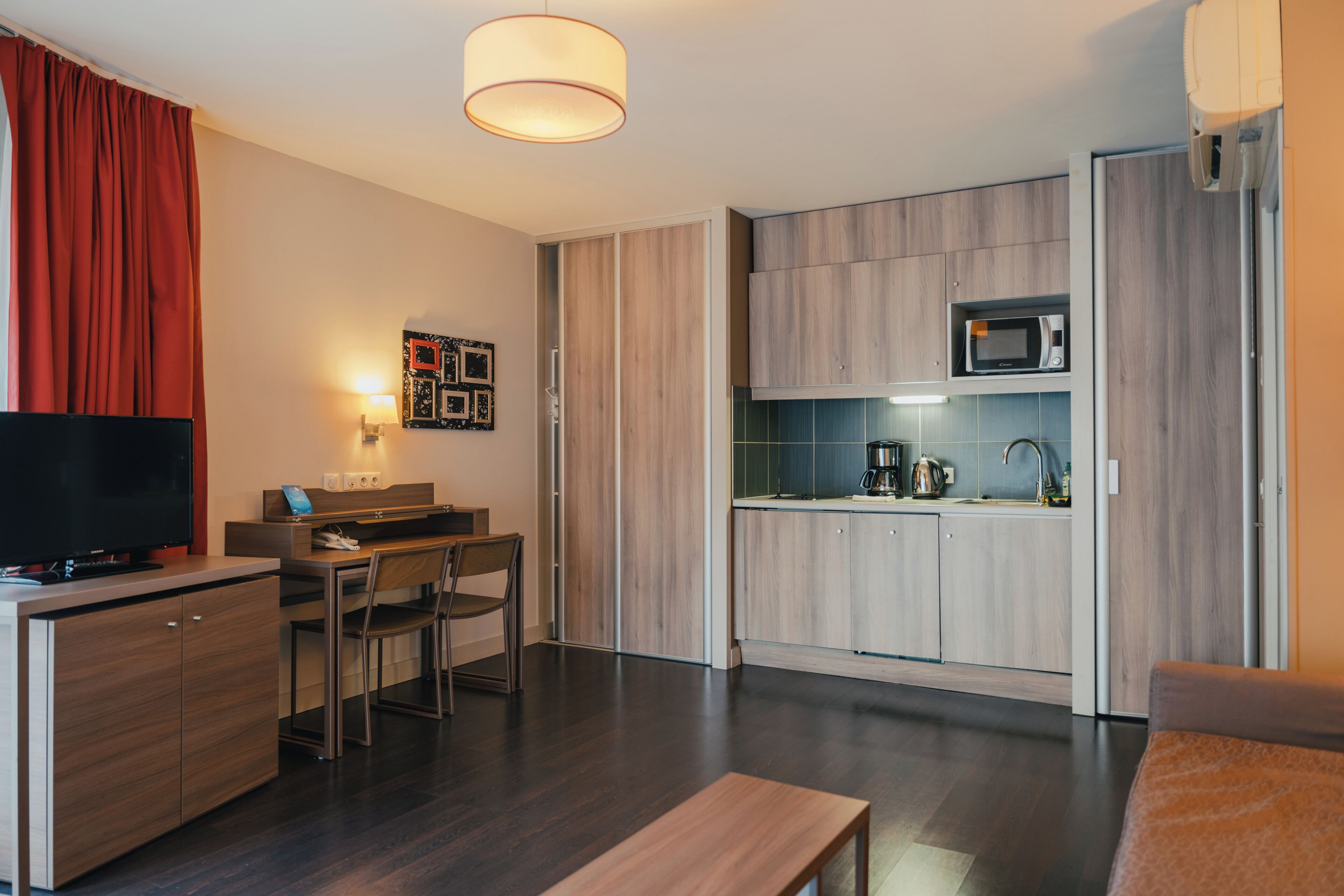A modern hotel room with a flat-screen TV, a desk, a chair, a small dining table, a microwave, and a coffee maker.