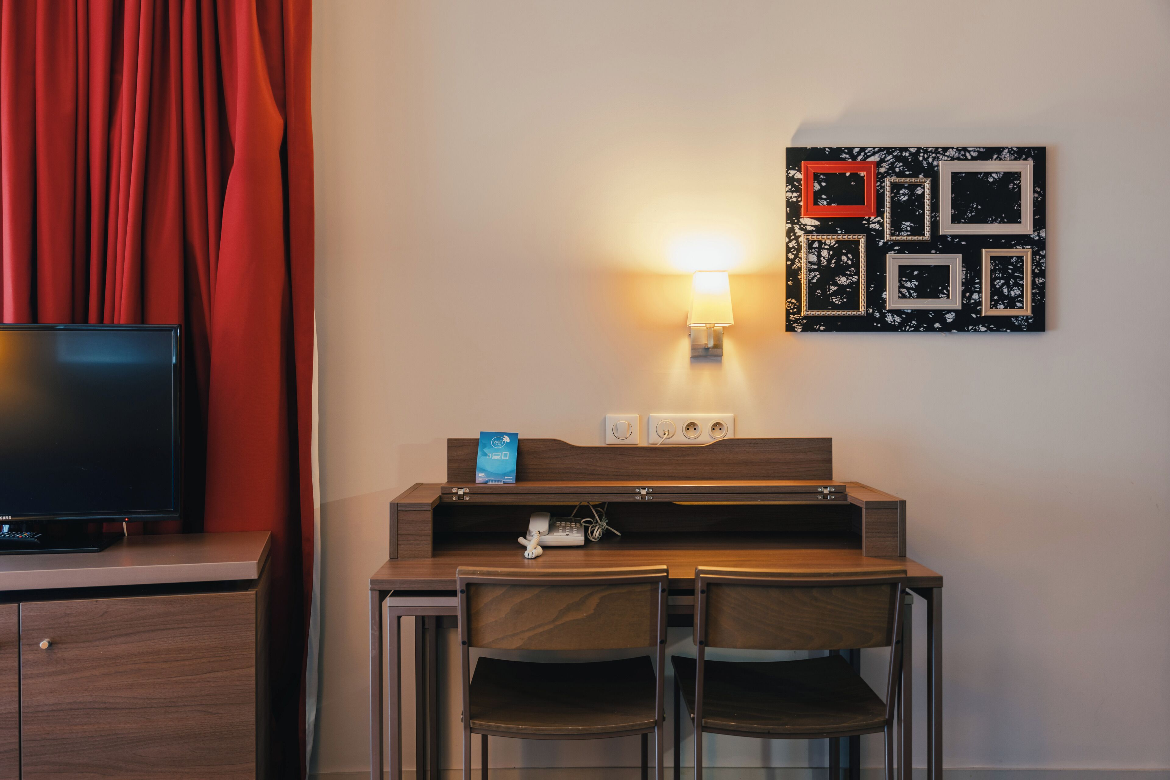 A hotel room with a TV, a desk with a lamp, a phone, and a wall-mounted picture frame.