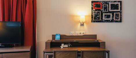 A hotel room with a TV, a desk with a lamp, a phone, and a wall-mounted picture frame.