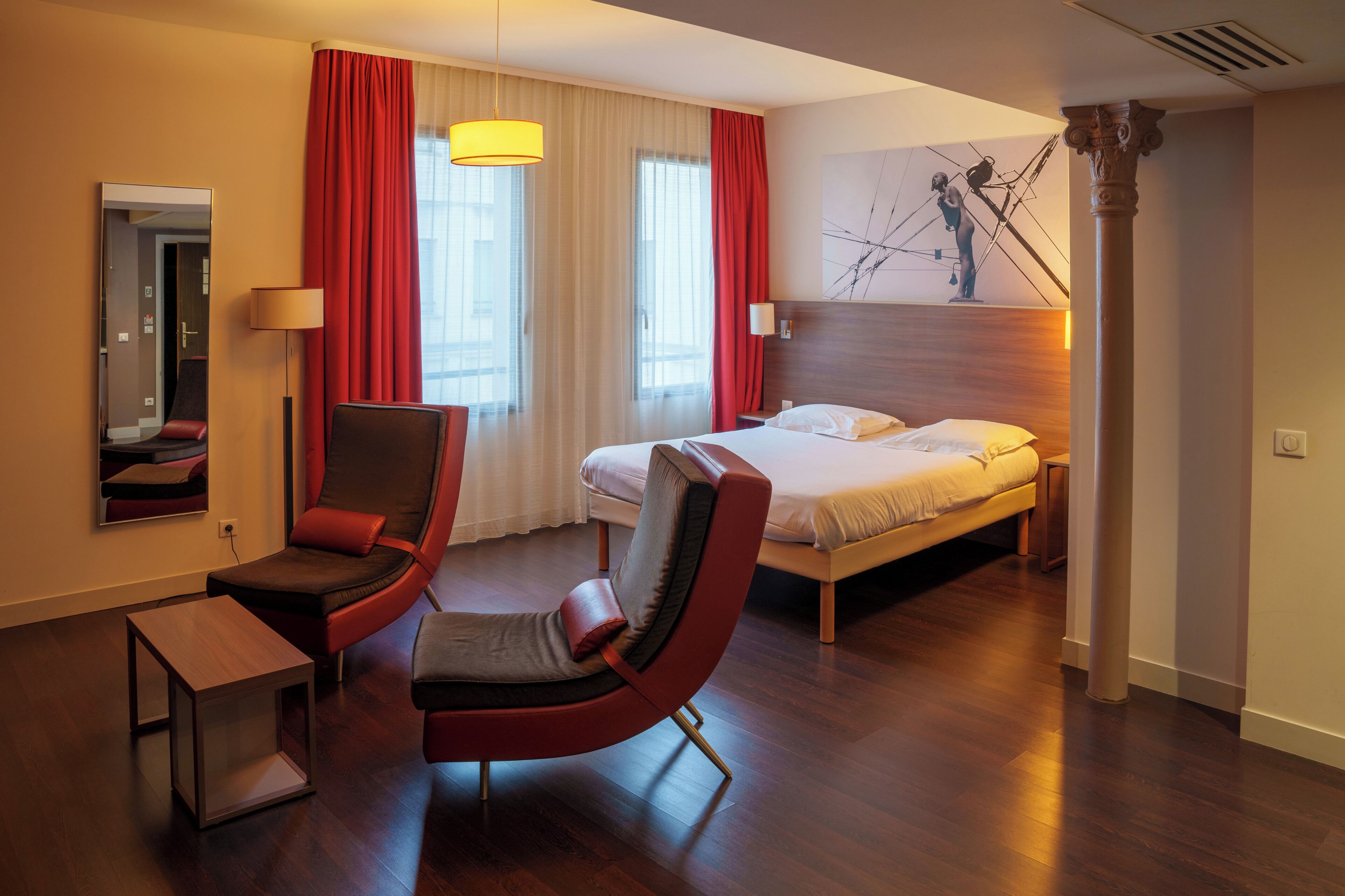 A hotel room with a bed, two chairs, a desk, a lamp, and a painting on the wall.