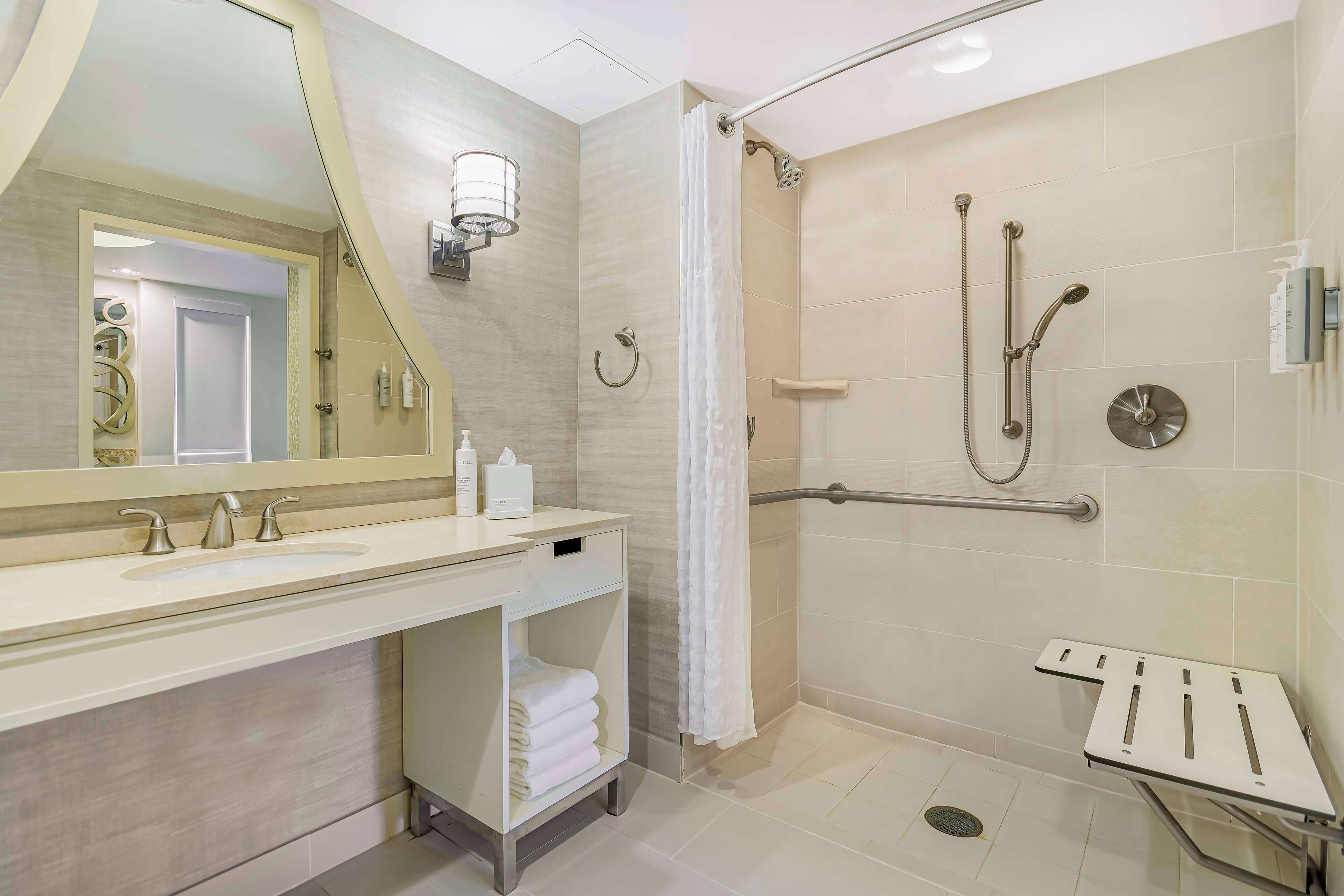 A modern bathroom with a large mirror, two sinks, a shower with a grab bar, and a towel rack.
