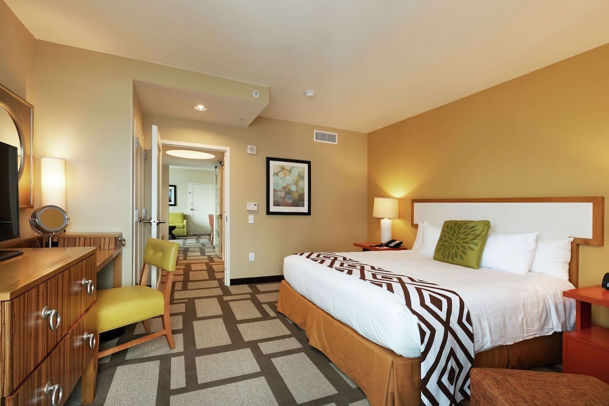 A hotel room with a bed, a desk with a mirror, a chair, a bedside table with a lamp, and a hallway leading to another room.