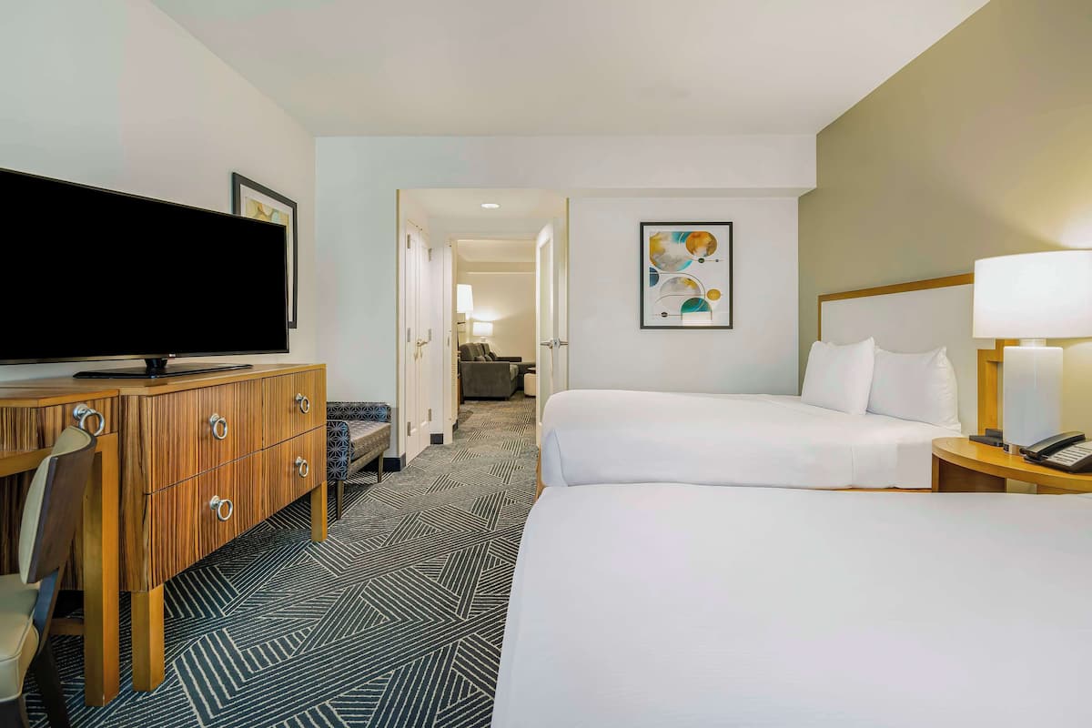A hotel room with a large flat-screen TV, a wooden dresser, two beds, a desk, and a lamp.