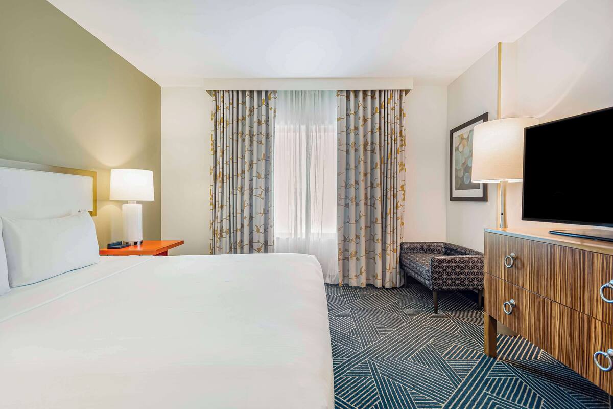 A hotel room with a large bed, a wooden dresser, a television, and a patterned chair.