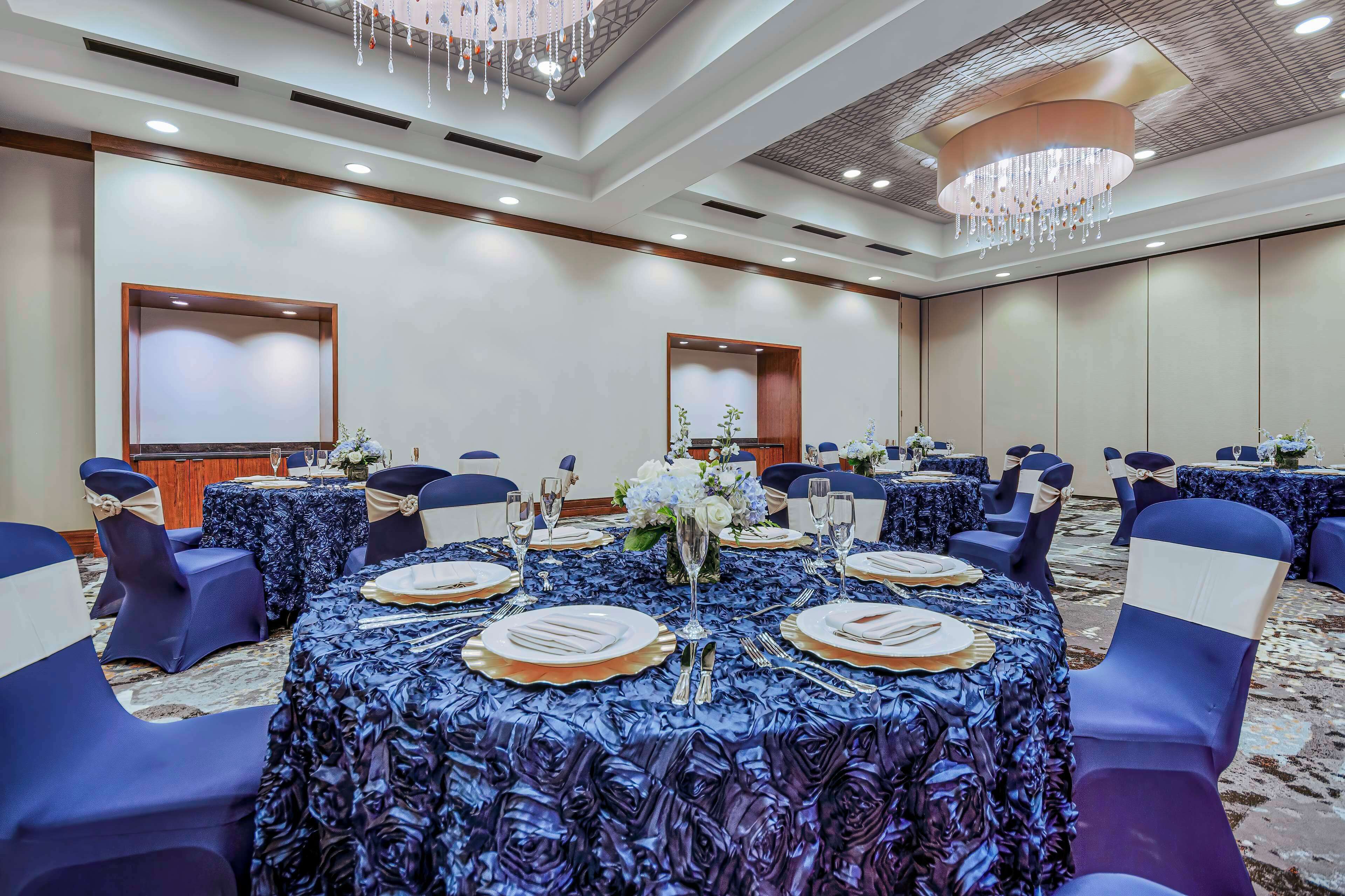 Une salle de banquet avec des tables rondes dressées pour le repas, chacune agrémentée d’assiettes blanches, de couverts et de verres.