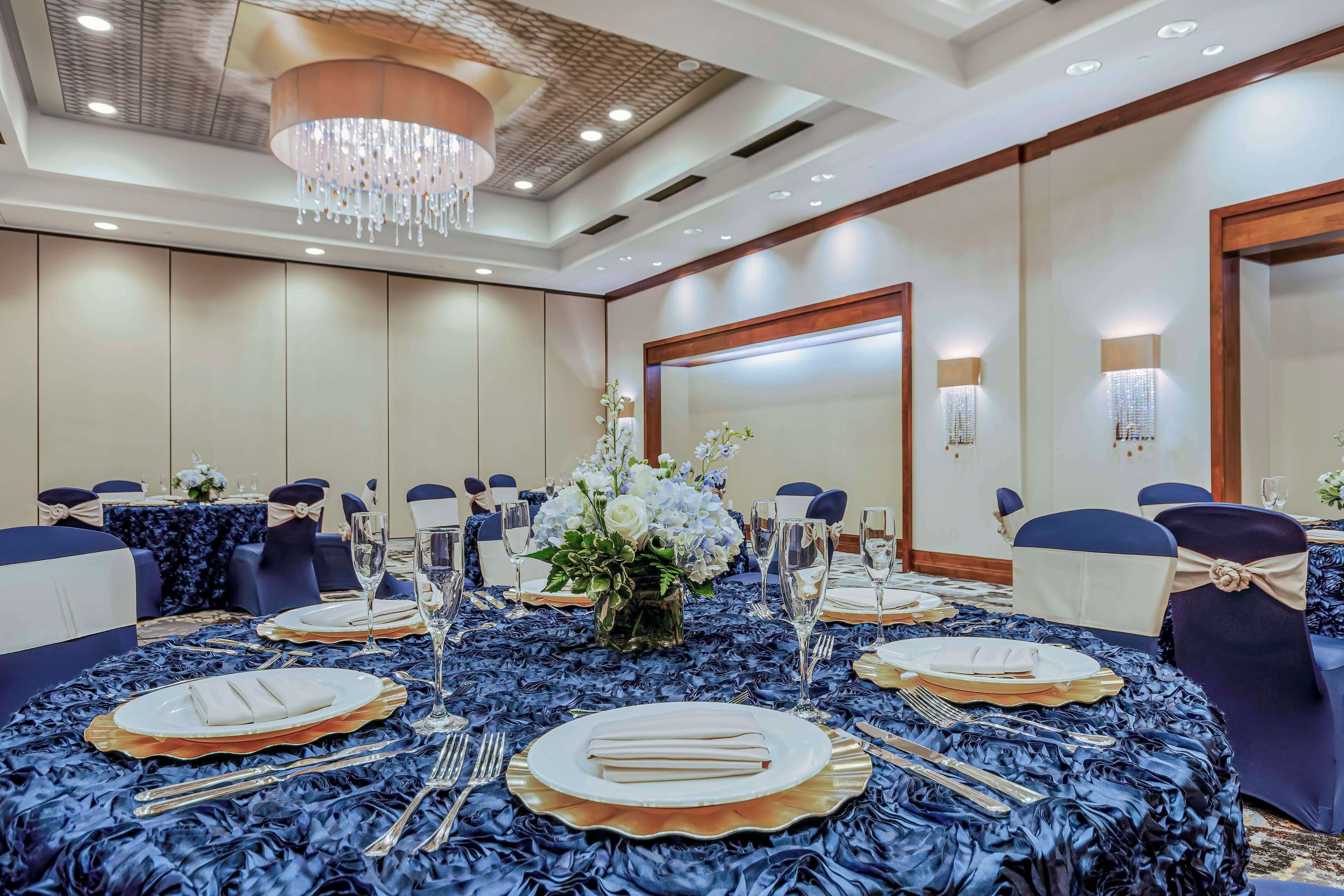 Un salón de conferencias con mesas redondas dispuestas para un evento formal, destacando un arreglo floral central, manteles azules y sillas a juego.