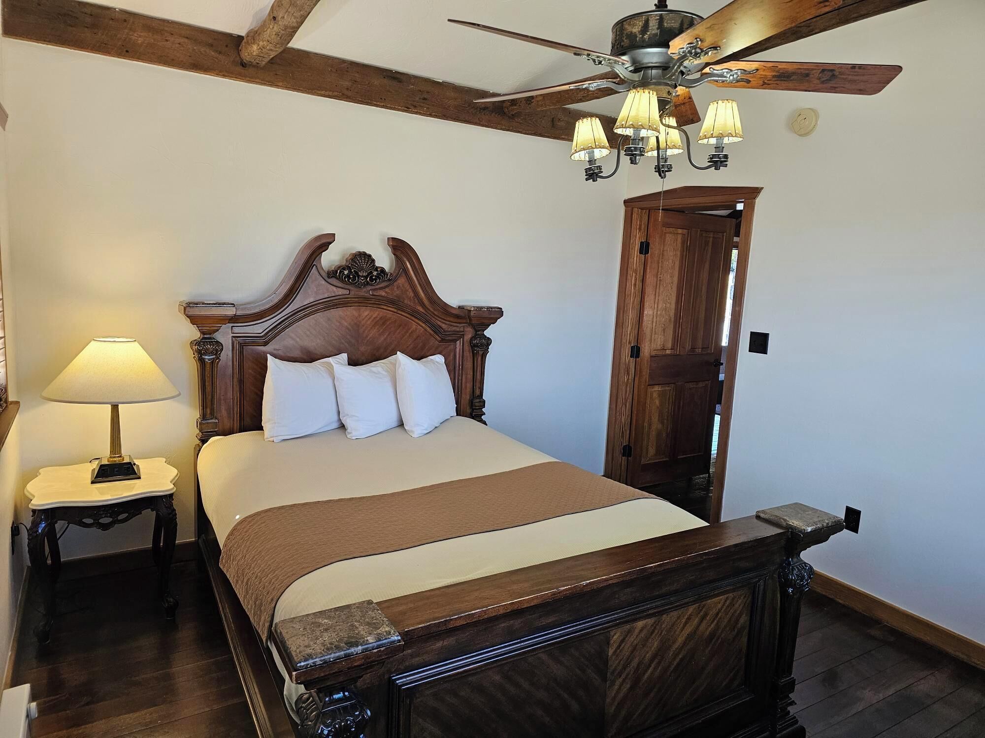 A bedroom with a wooden bed, a nightstand with a lamp, a ceiling fan, and a wooden door.