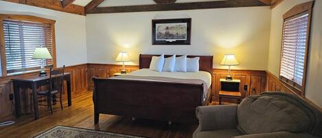 A bedroom with a large bed, wooden headboard, two bedside tables with lamps, a desk with a chair, and a patterned rug.