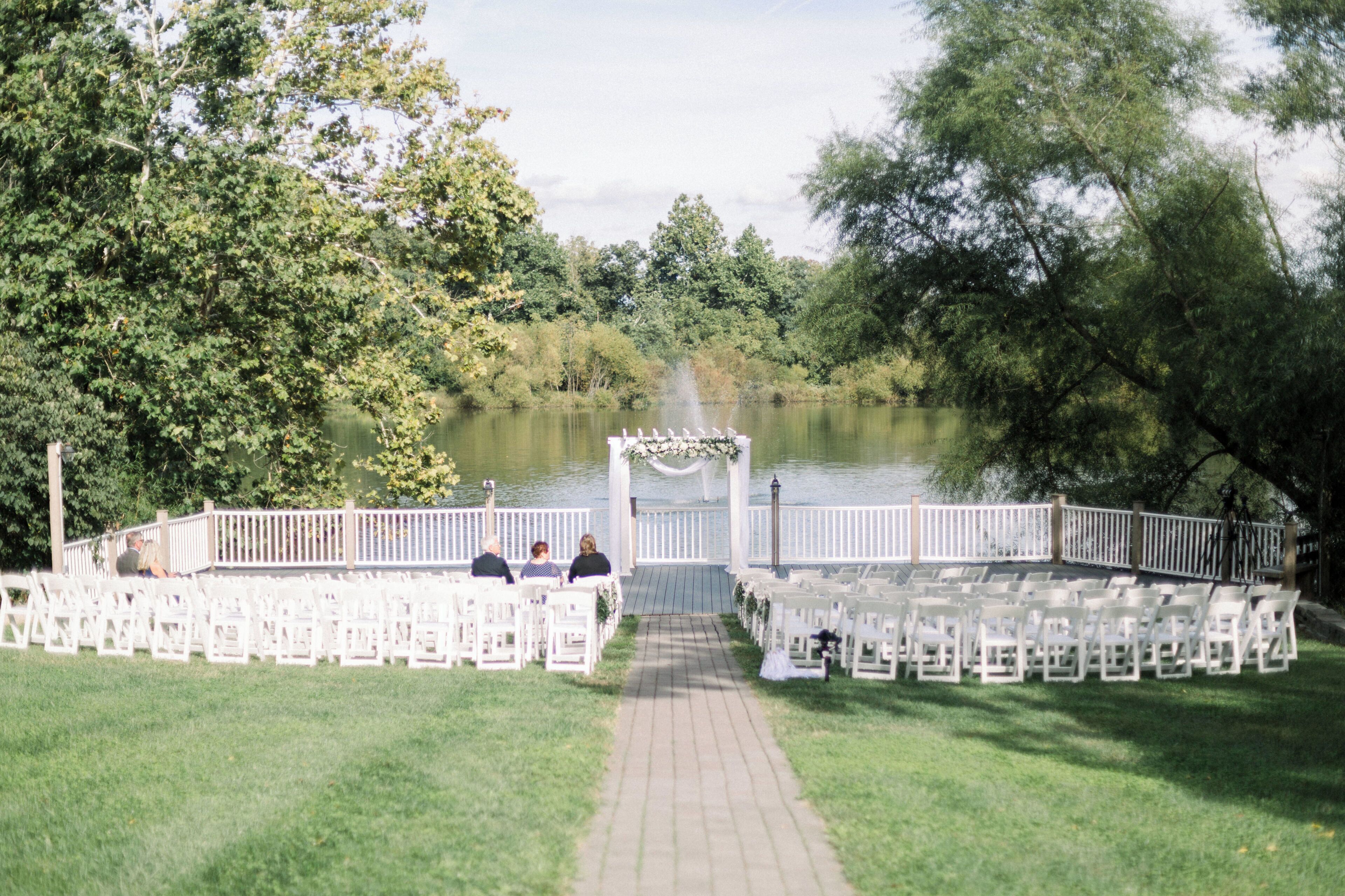 A lakeside outdoor wedding ceremony with white chairs arranged in rows, a white archway, and a wooden deck.