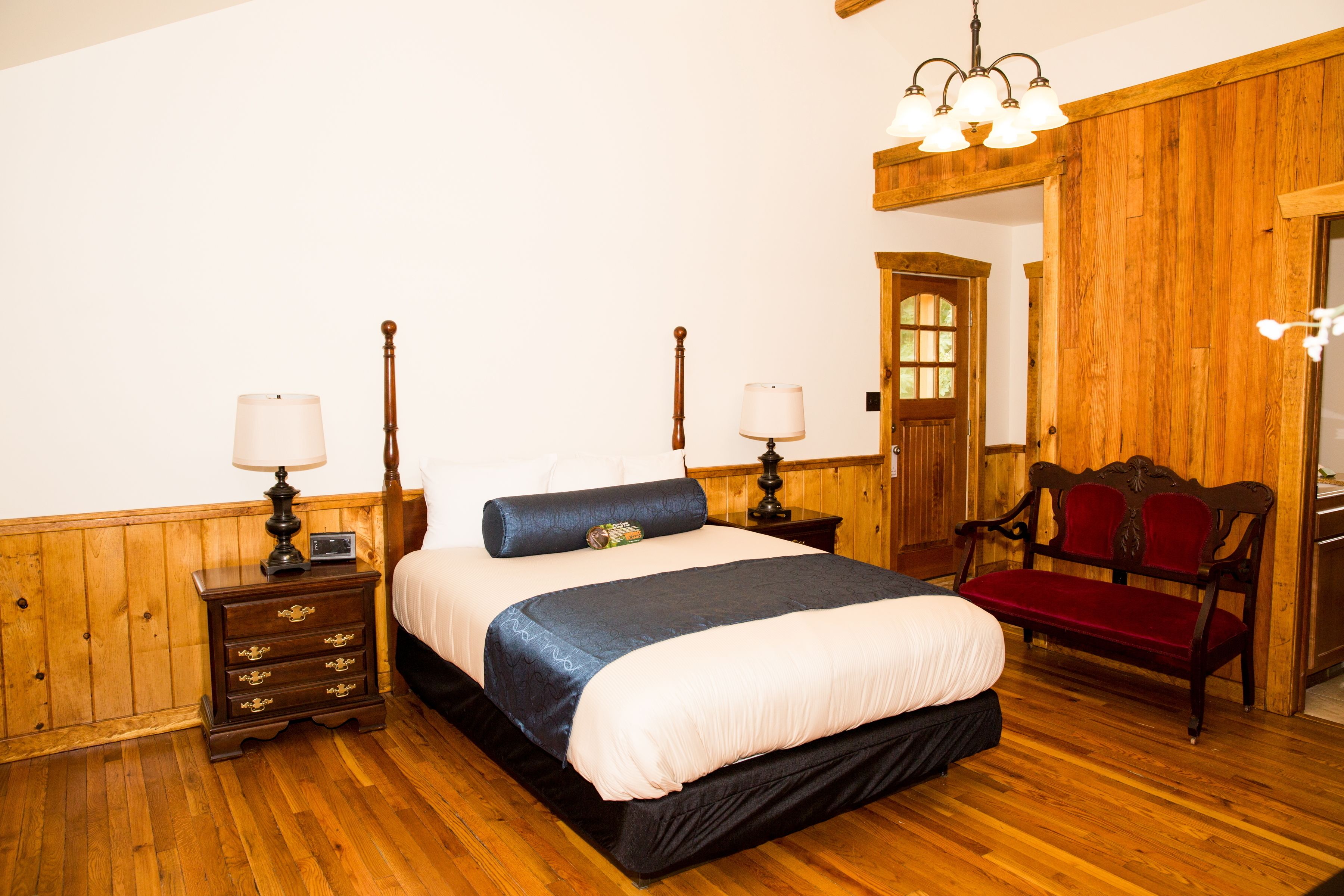 A bedroom with a wooden floor, a bed with a blue and white bedspread, two bedside tables with lamps, a red bench, and a wooden door.