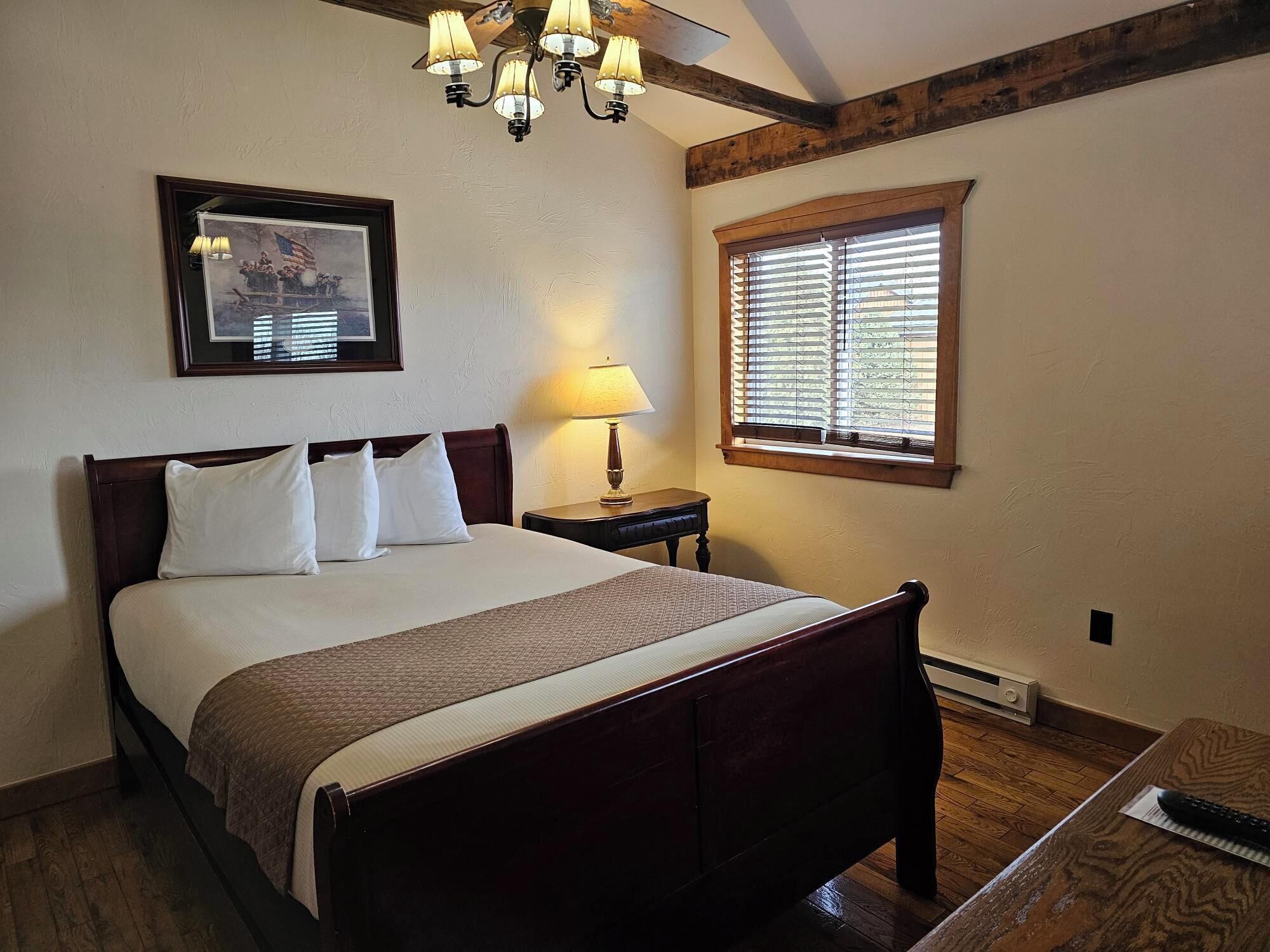 Un dormitorio con cama de madera, una mesita de noche, una lámpara, un cuadro enmarco y una ventana con persianas.
