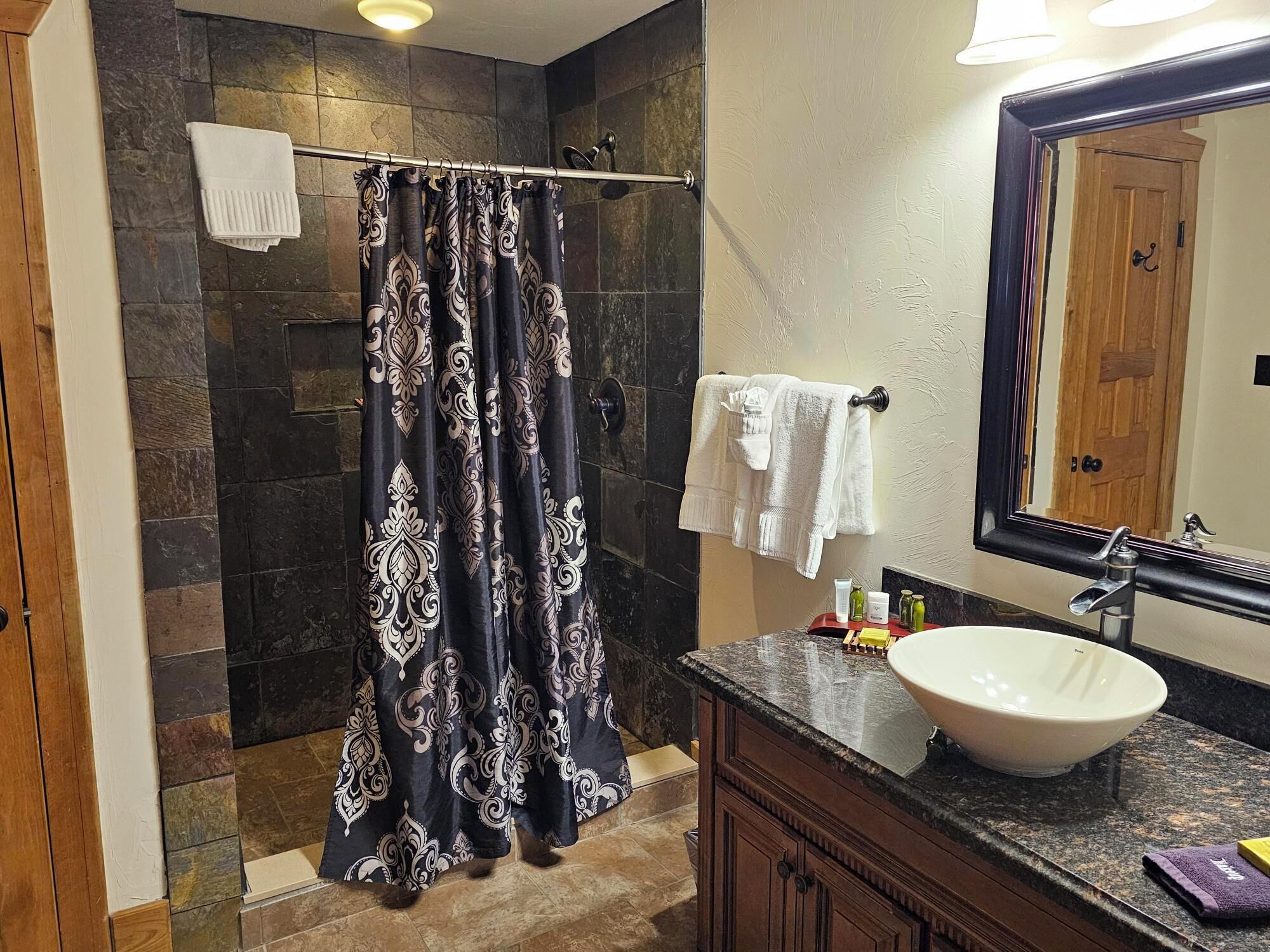 A bathroom with a stone shower wall, a black and white patterned shower curtain, a white towel, a large mirror, and a sink with toiletries.