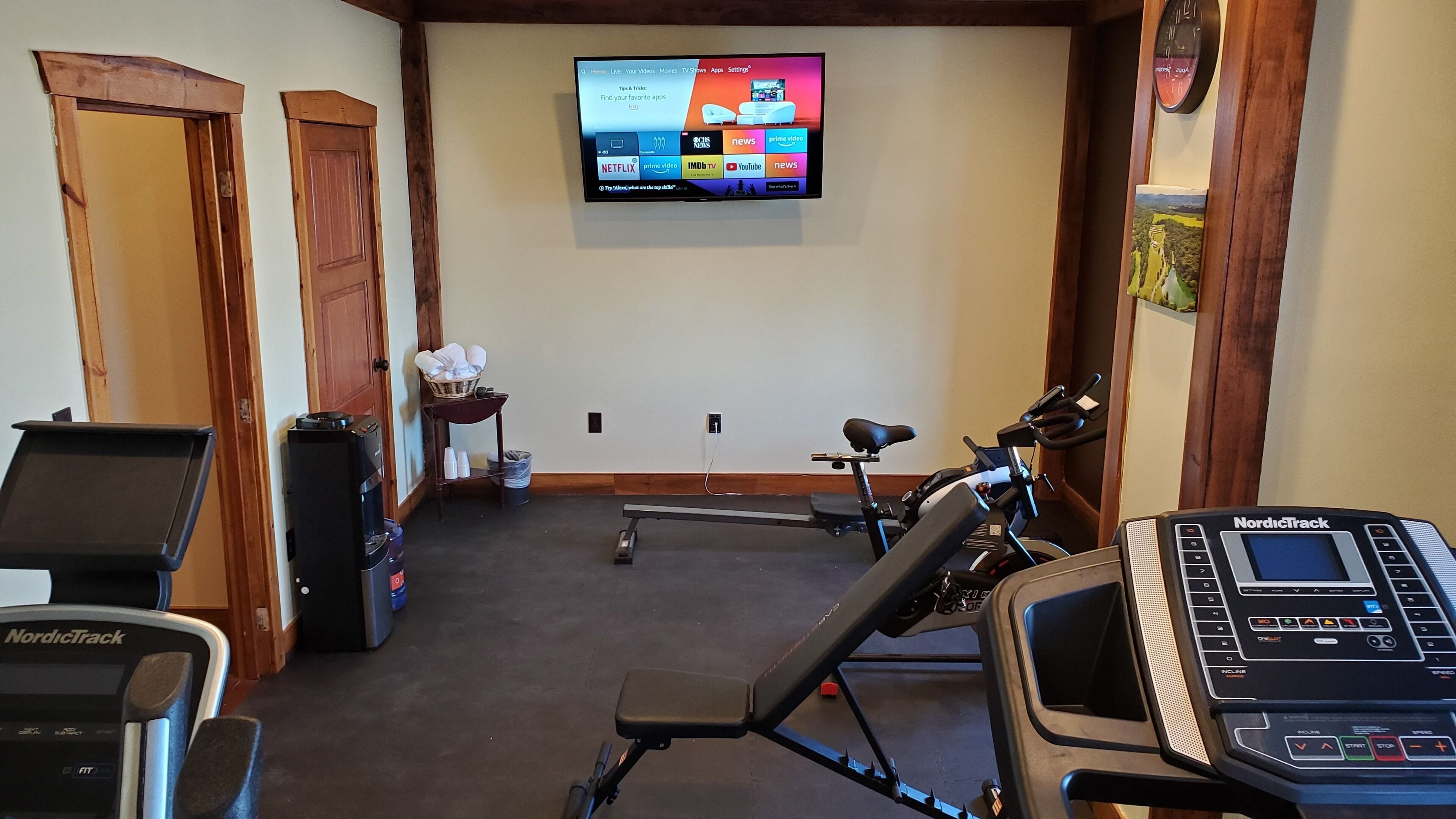 A home gym with treadmills, an elliptical, and a NordicTrack. A TV mounted on the wall displays various apps.