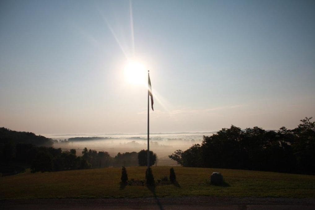 A flagpole in a field with a clear sky and sun in the background.