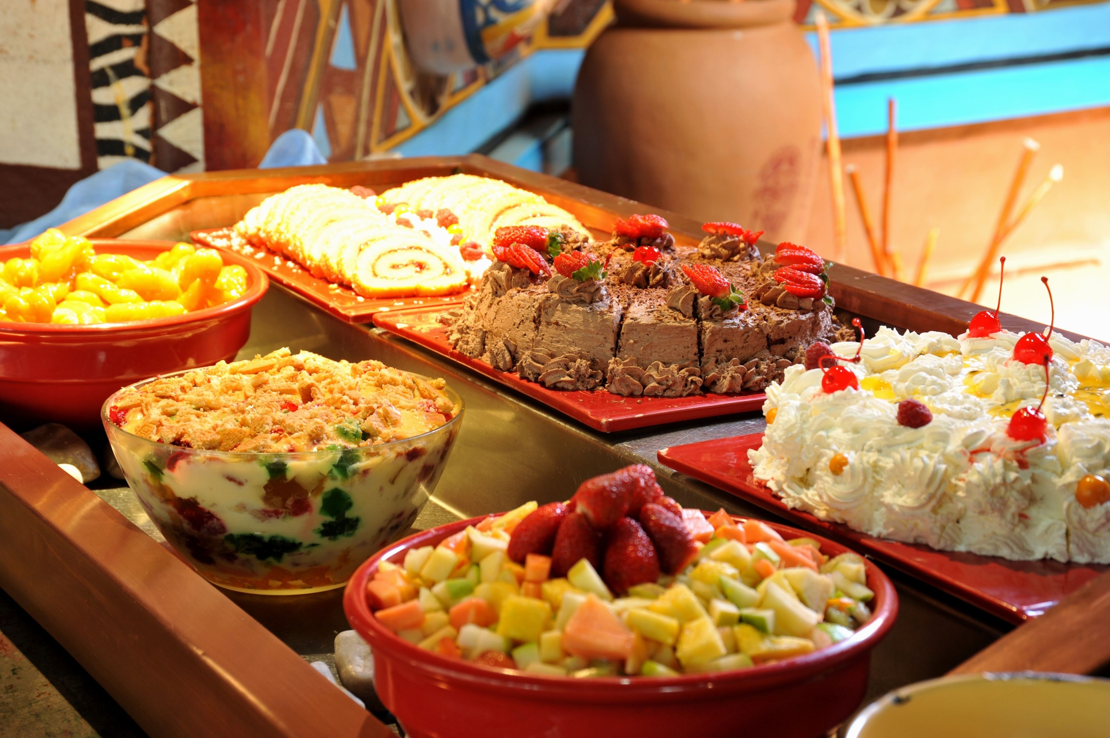 Taula de buffet amb diversos plats, incloent-hi una amanida de fruita, un pastís de xocolata i un postre amb nata i cireres.