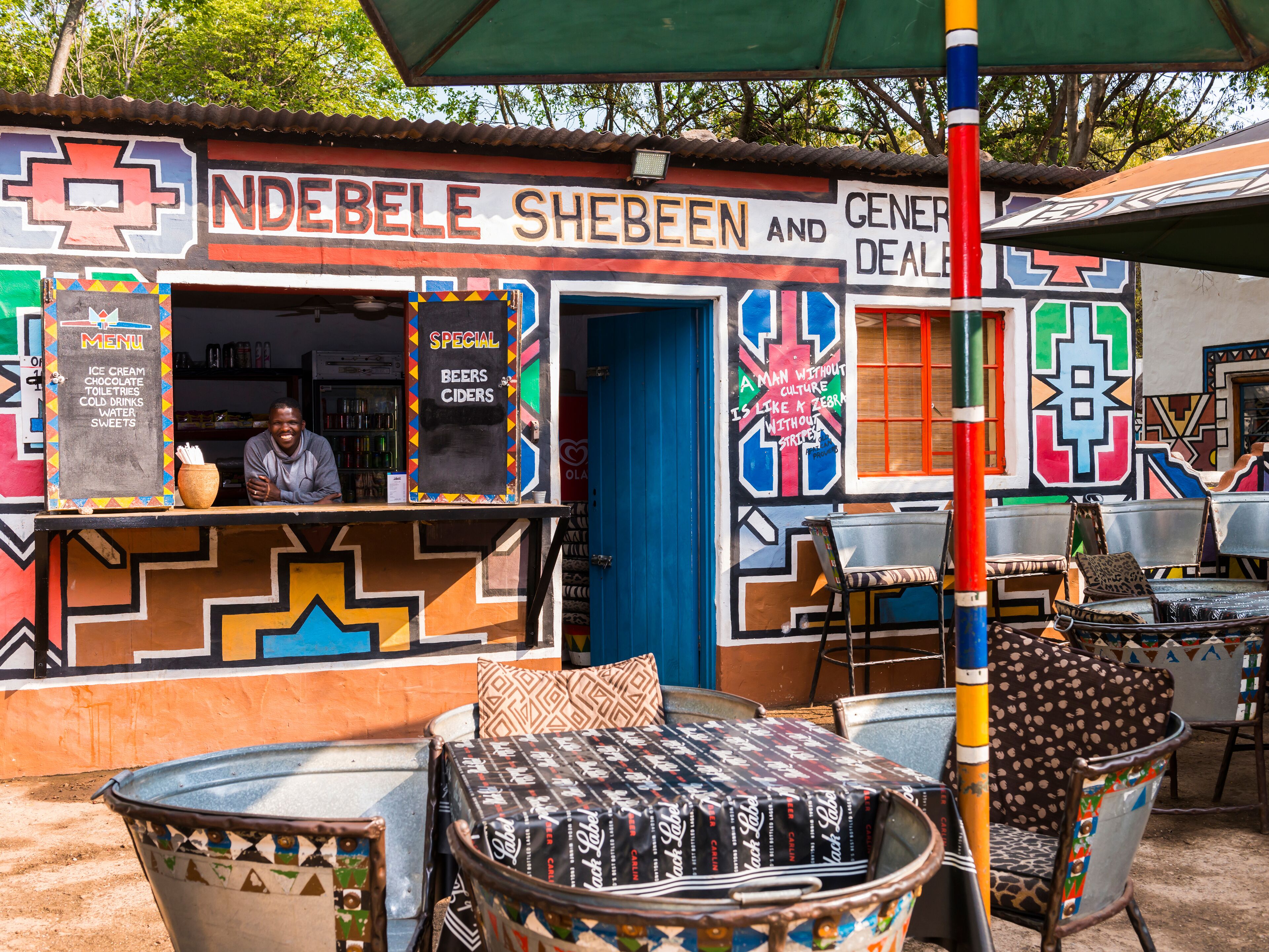 An outdoor seating area with colorful chairs and tables, a chalkboard menu, and a building with vibrant murals.