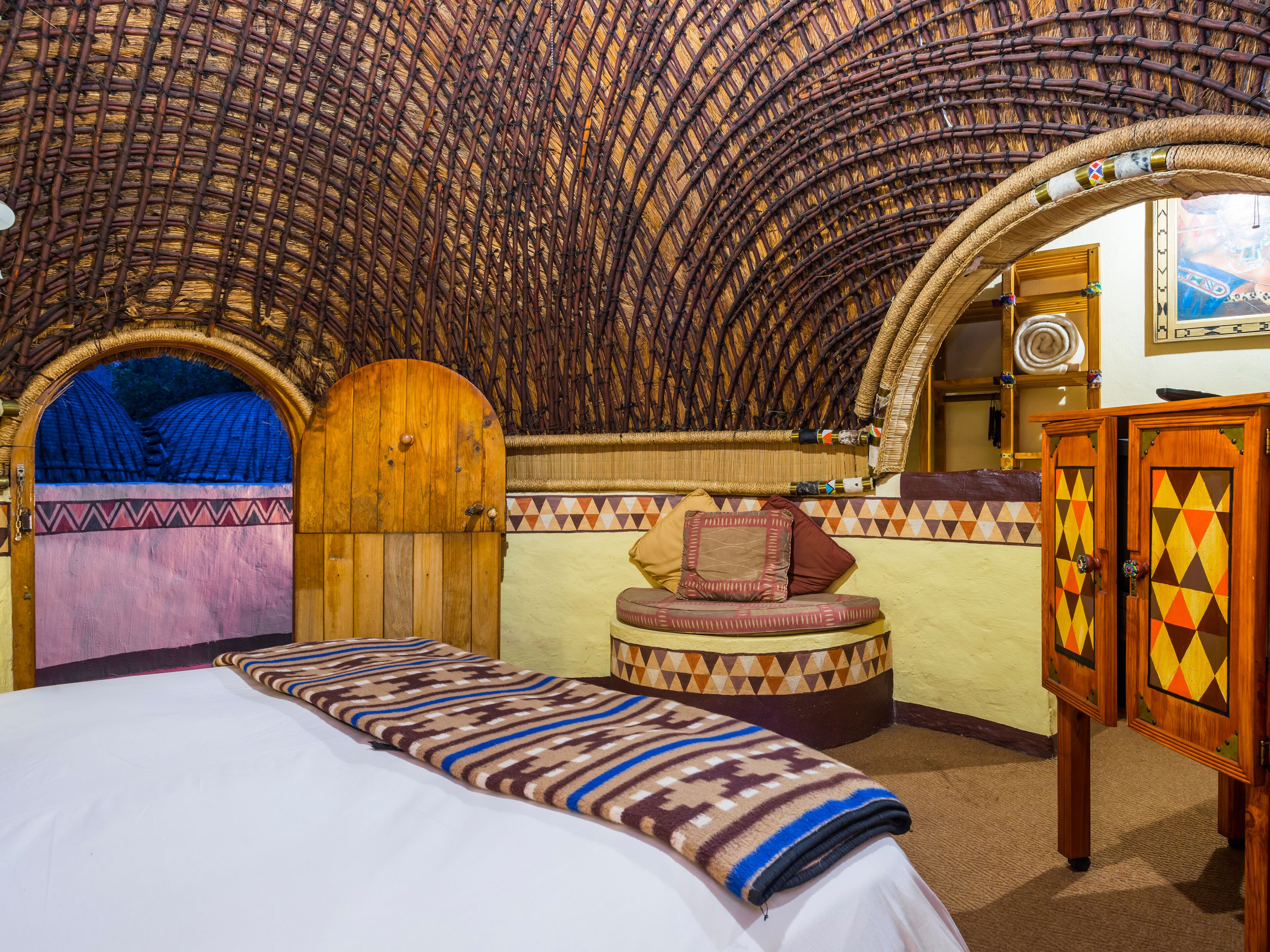 A room with a high, woven ceiling, a bed with a patterned blanket, a wooden door, and a small seating area with cushions.
