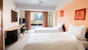 A hotel room with two beds, a desk with a chair, a television, and a view of buildings through large windows. - Beer Garden Inn (Addis Ababa)