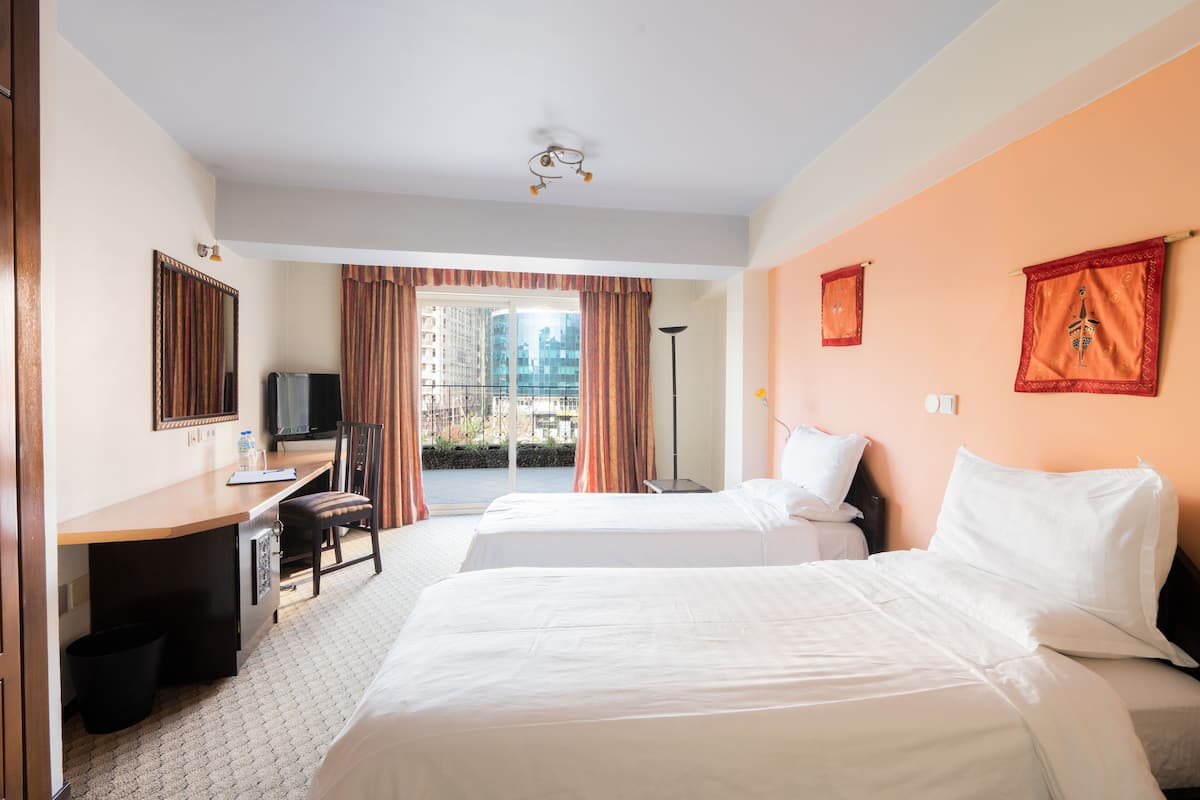 A hotel room with two beds, a desk with a chair, a television, and a view of buildings through large windows.