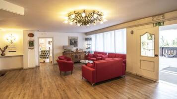 Hall d'albergo con un divano rosso, un tavolino da caffè e un lampadario. Si nota la reception con una segnaletica per bar e ristorante.