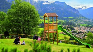 A playground with a wooden climbing structure and a green slide, surrounded by lush greenery and a mountainous backdrop. - Centro Vacanze Veronza Clubresidence (Ville di Fiemme)