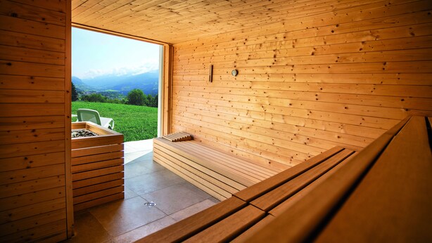 A wooden sauna with a bench and a small table, a window with a view of mountains, and a wooden wall. - Centro Vacanze Veronza Clubresidence (Ville di Fiemme)