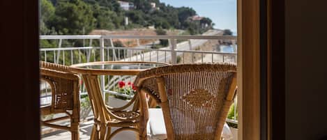 A balcony with wicker chairs and a table, overlooking a coastal landscape with buildings and greenery.