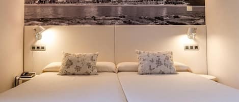 Una habitación de hotel con una cama grande, dos almohadas, lámparas de noche, una mesita de noche con un teléfono y un cuadro colgado en la pared que muestra un paisaje costero.