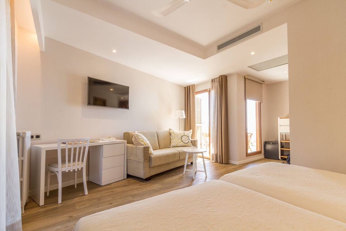 Habitación de hotel con cama, escritorio con silla, sofá, televisor montado en la pared y ventana con cortinas.