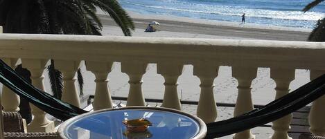 Un balcón con una mesa de vidrio, sillas de mimbre y vistas a la playa y al océano.