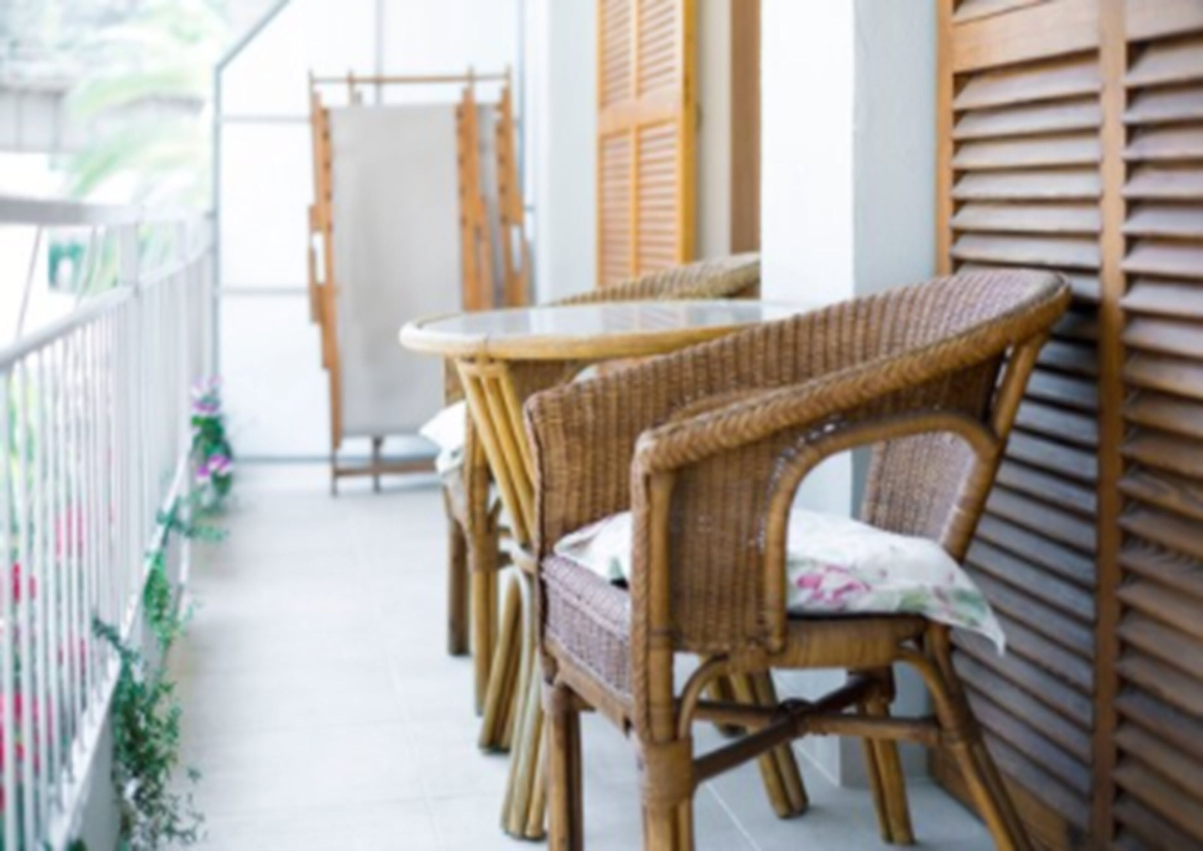 Un balcón con sillas de mimbre, una mesa, barandilla blanca y pared con contraventanas de madera.
