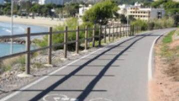 Un sendero pavimentado con indicaciones para bicicletas, delimitado por una valla, que conduce a una playa y montañas.
