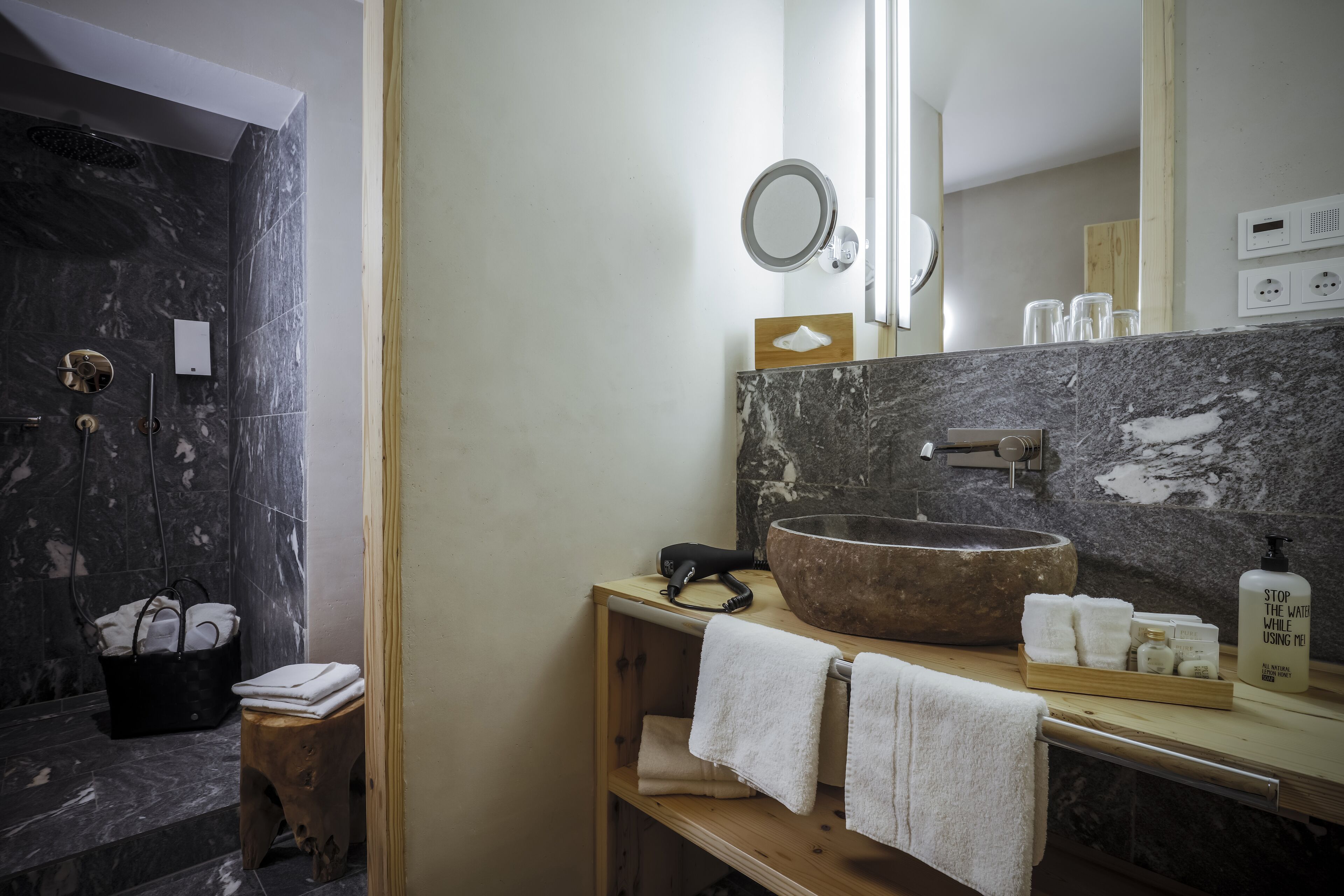 A modern bathroom with a large stone sink, wooden vanity, and a shower area with dark marble tiles.