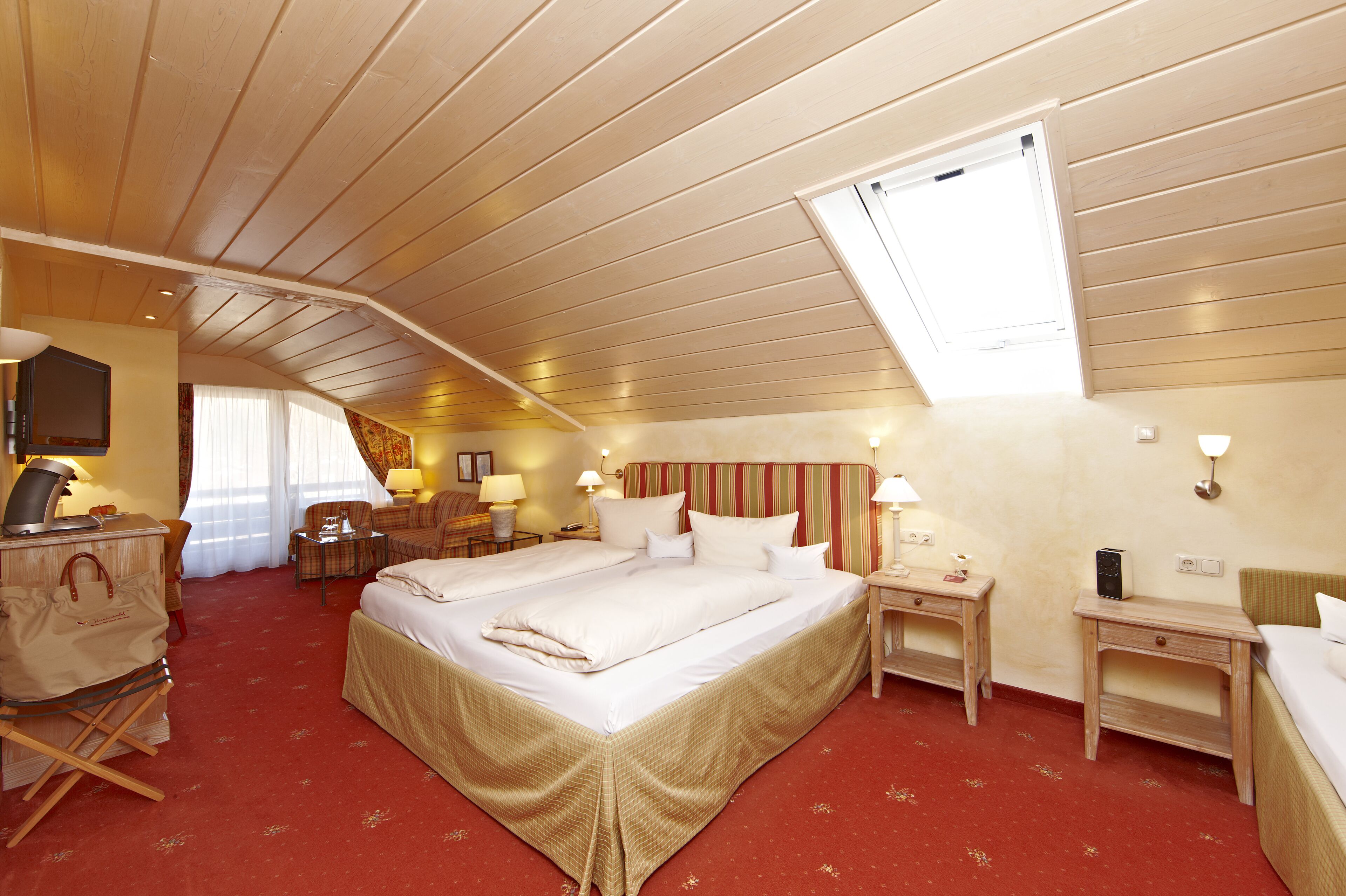 A hotel room with a large bed, a desk, a chair, a small table, a TV, and a window with a view of the sky.