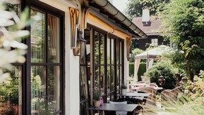 An outdoor patio with tables and chairs, large windows, and potted plants. - Staudacherhof (Garmisch-Partenkirchen)