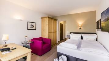 A hotel room with a bed, a desk with a lamp, a chair, a wardrobe, and a small table with a lamp.