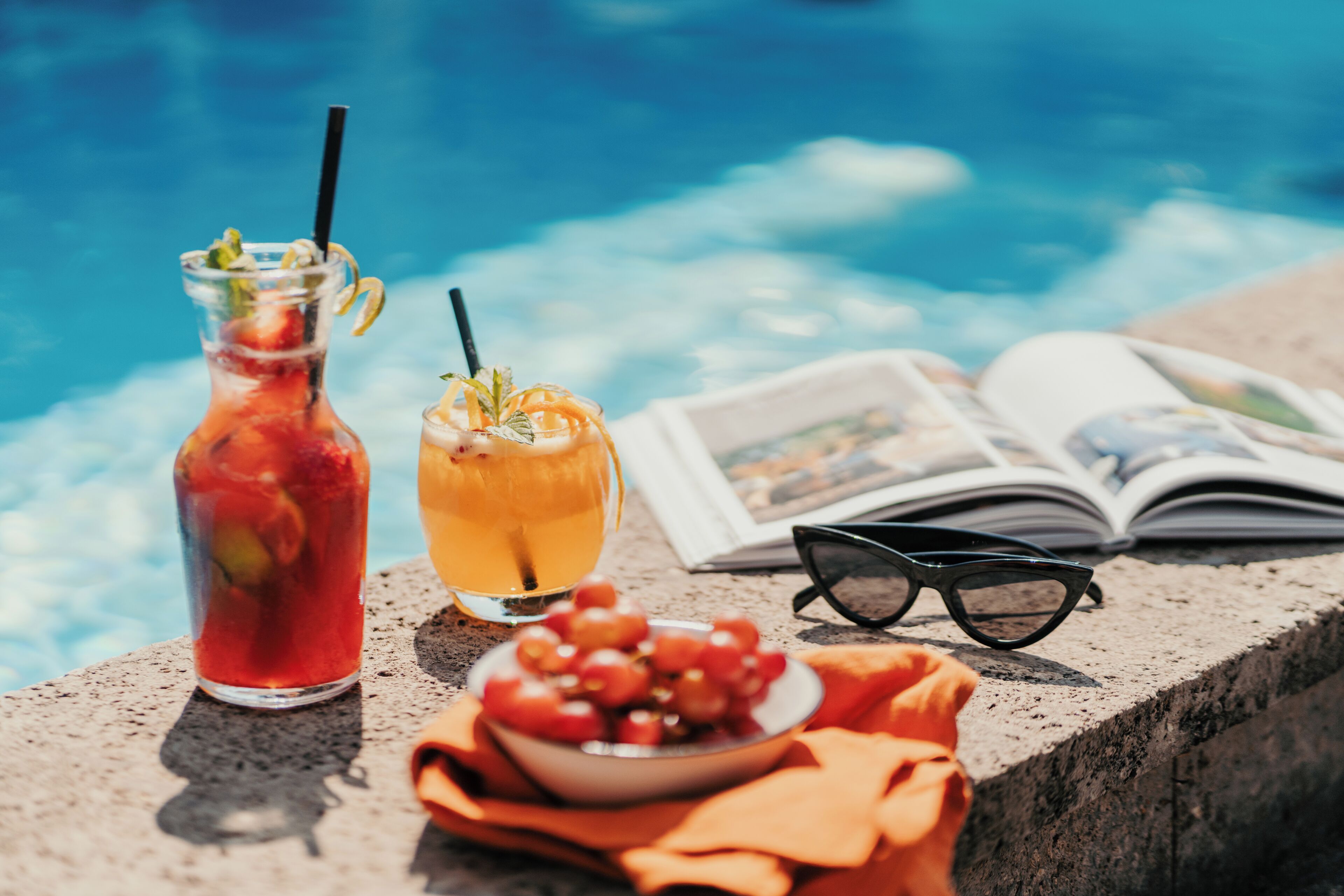 A glass bottle, a glass with a straw, a bowl of grapes, a pair of glasses, and a book by a pool.