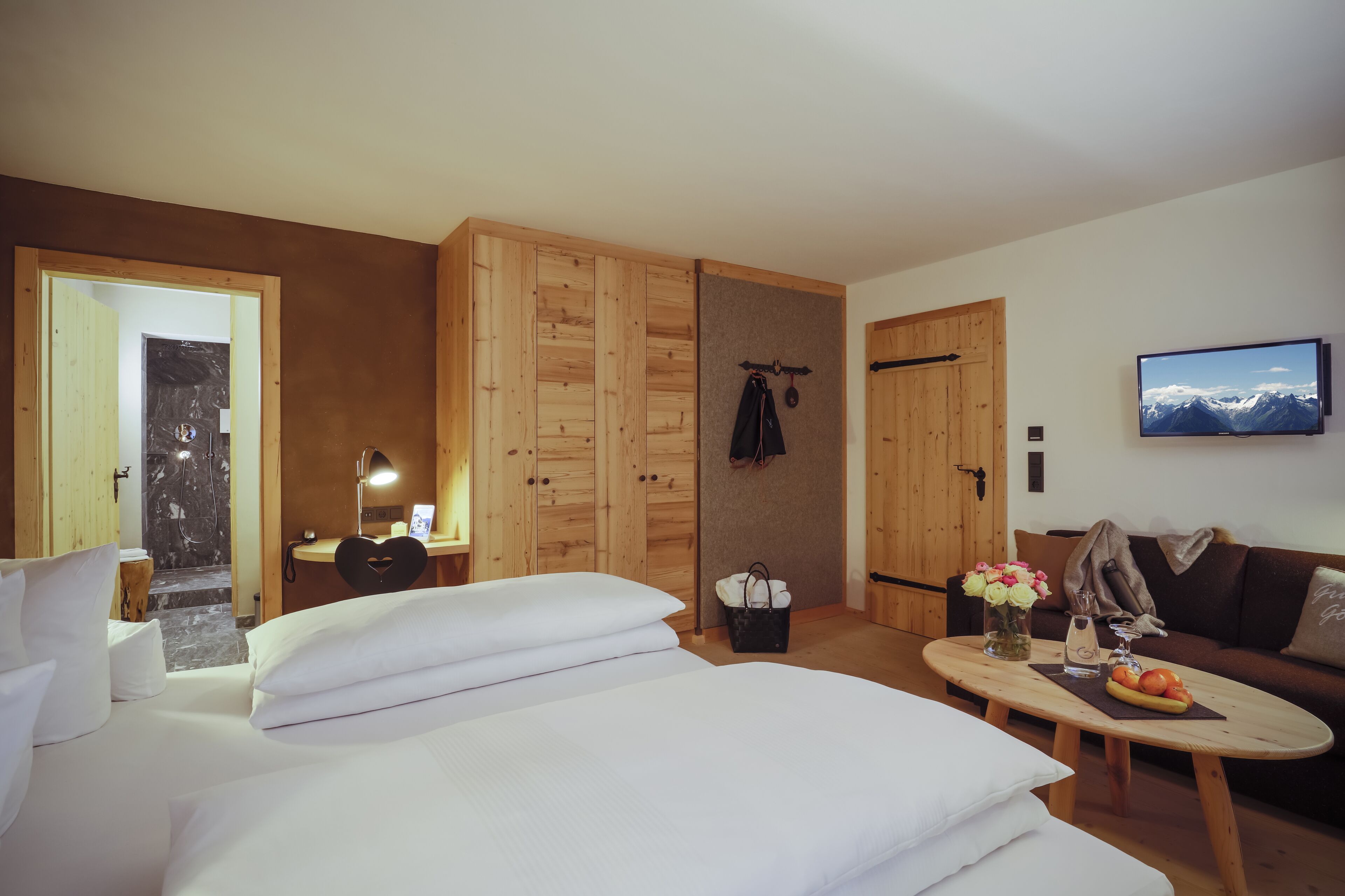 A hotel room with a bed, a wooden wardrobe, a small desk with a lamp, a television, and a coffee table with fruits and a vase of flowers.
