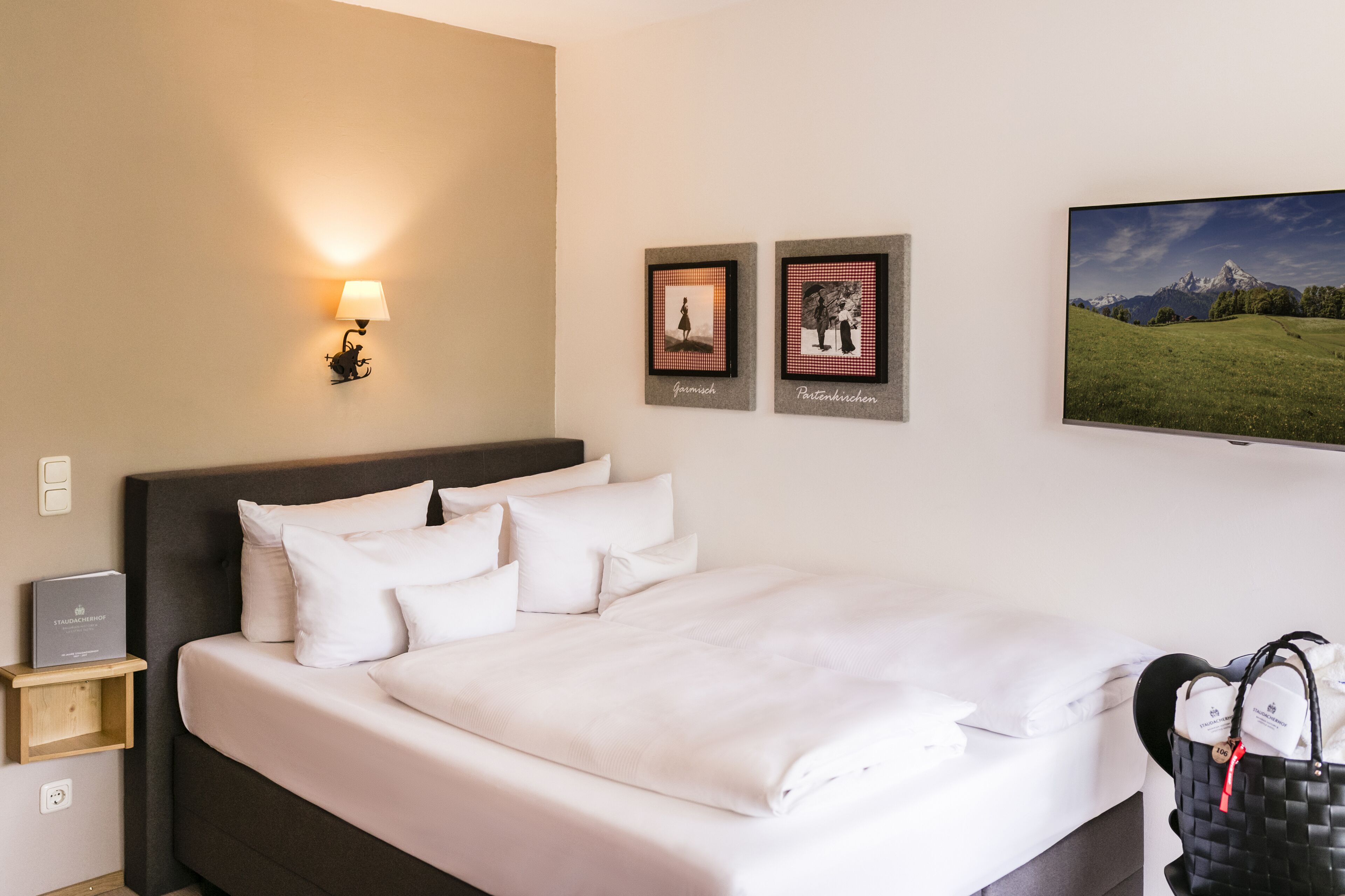 A neatly made bed with white linens, a wall-mounted lamp, two framed pictures, and a television on the wall.