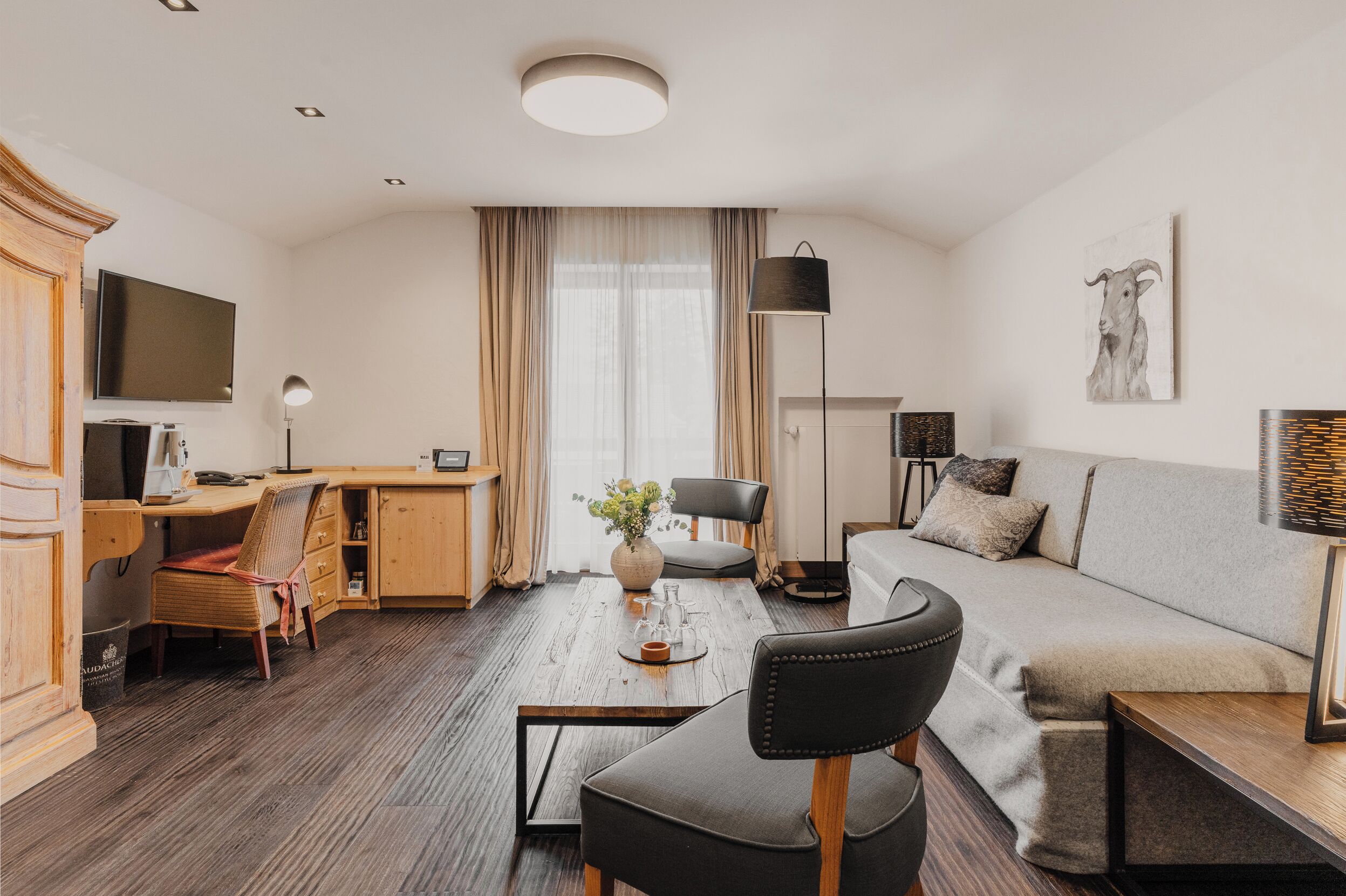 A modern hotel room with a wooden desk, a sofa, a TV, and a large window with curtains.