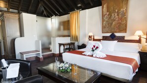 A hotel room with a bed, a desk, a chair, a small table with a vase of flowers, and a painting on the wall. - Casa Palacio Pilar del Toro (Granada)