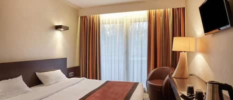 A hotel room with a bed, a desk with a lamp, a television, and a window with curtains.