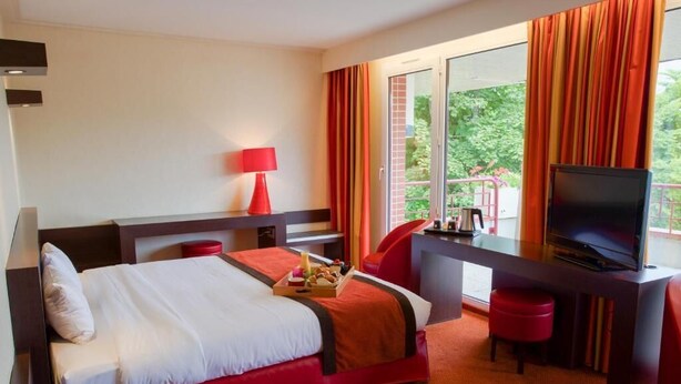 Een hotelkamer met een bed, een bureau met een televisie, een rode stoel en een raam met gordijnen.
