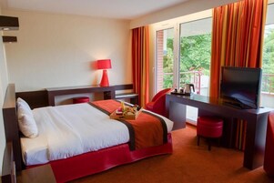 Habitación de hotel con cama, escritorio con televisor, una silla roja y ventana con cortinas.