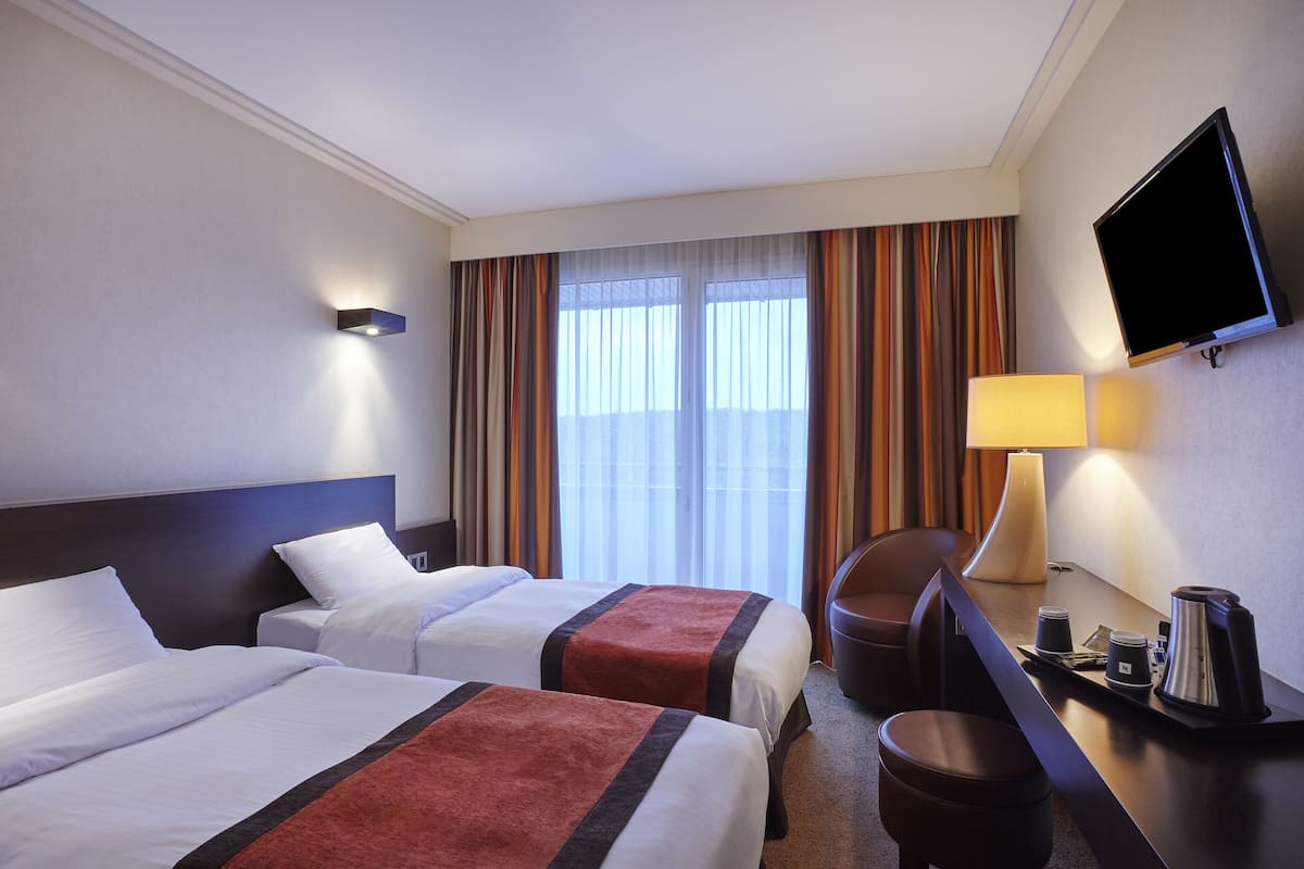 Een hotelkamer met twee bedden, een bureau met een lamp, een televisie en uitzicht op het omliggende landschap door grote ramen.