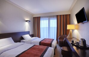 Een hotelkamer met twee bedden, een bureau met een lamp, een televisie en uitzicht op het omliggende landschap door grote ramen.